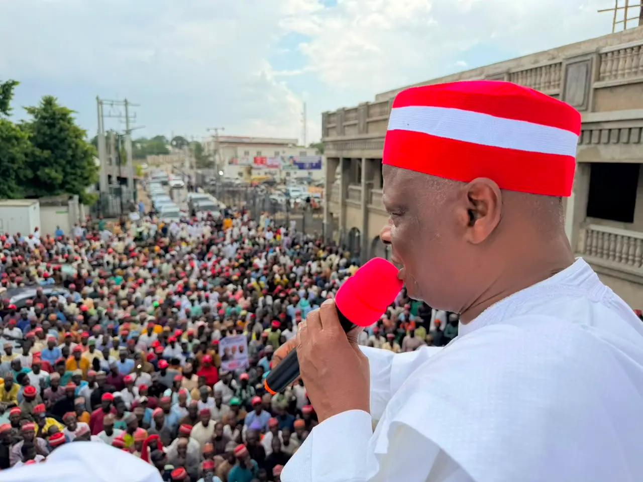 Rabiu Kwankwaso in a red and white cap speaking into a microphone from a balcony above a street packed with thousands of people