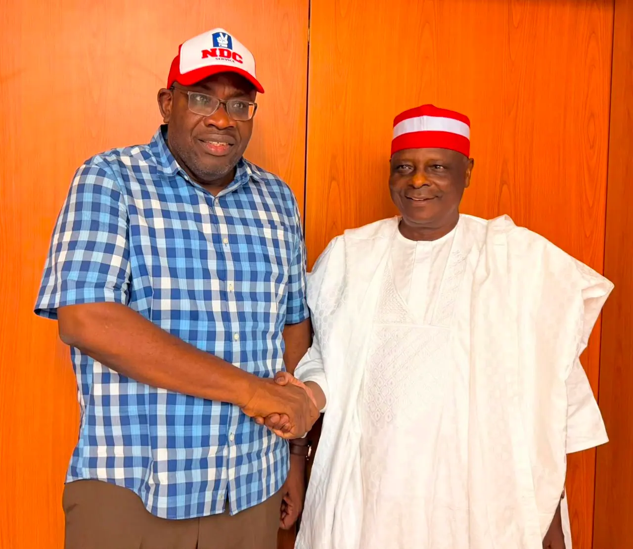Rabiu Kwankwaso shaking hands with Senator Seriake Dickson, who is wearing a Nigeria Democratic Congress cap, in front of a wooden wall