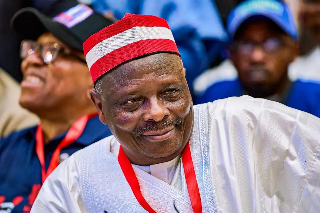 Rabiu Kwankwaso in a red and white cap, smiling, seated among delegates in dark blue caps and lanyards
