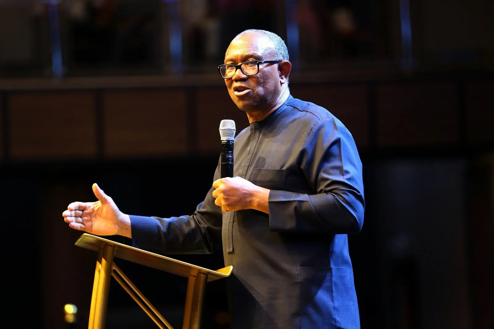 Peter Obi speaking into a handheld microphone with his other hand open, lit against a dark background