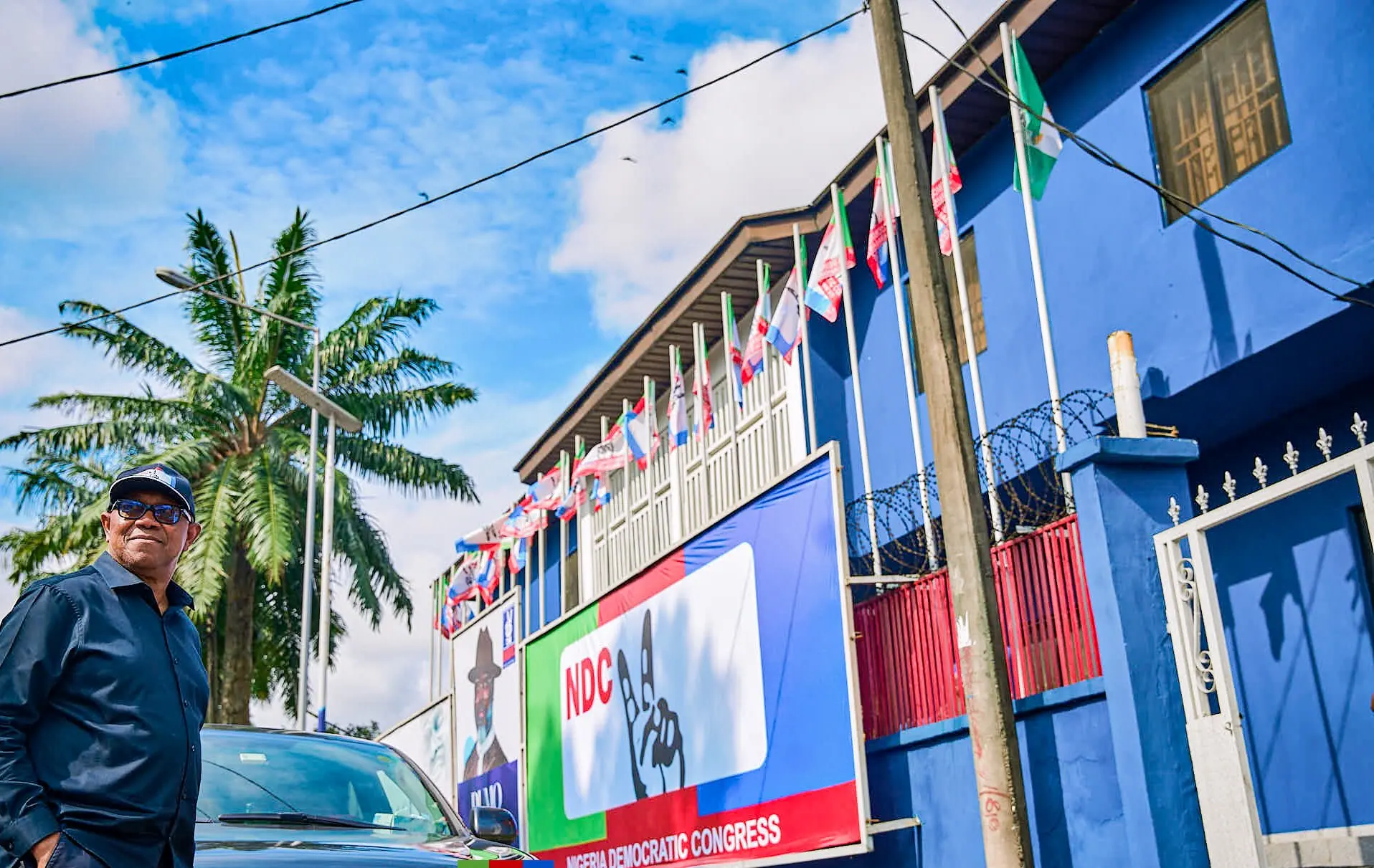Peter Obi standing beside a vehicle on a street of blue buildings lined with flags and palms