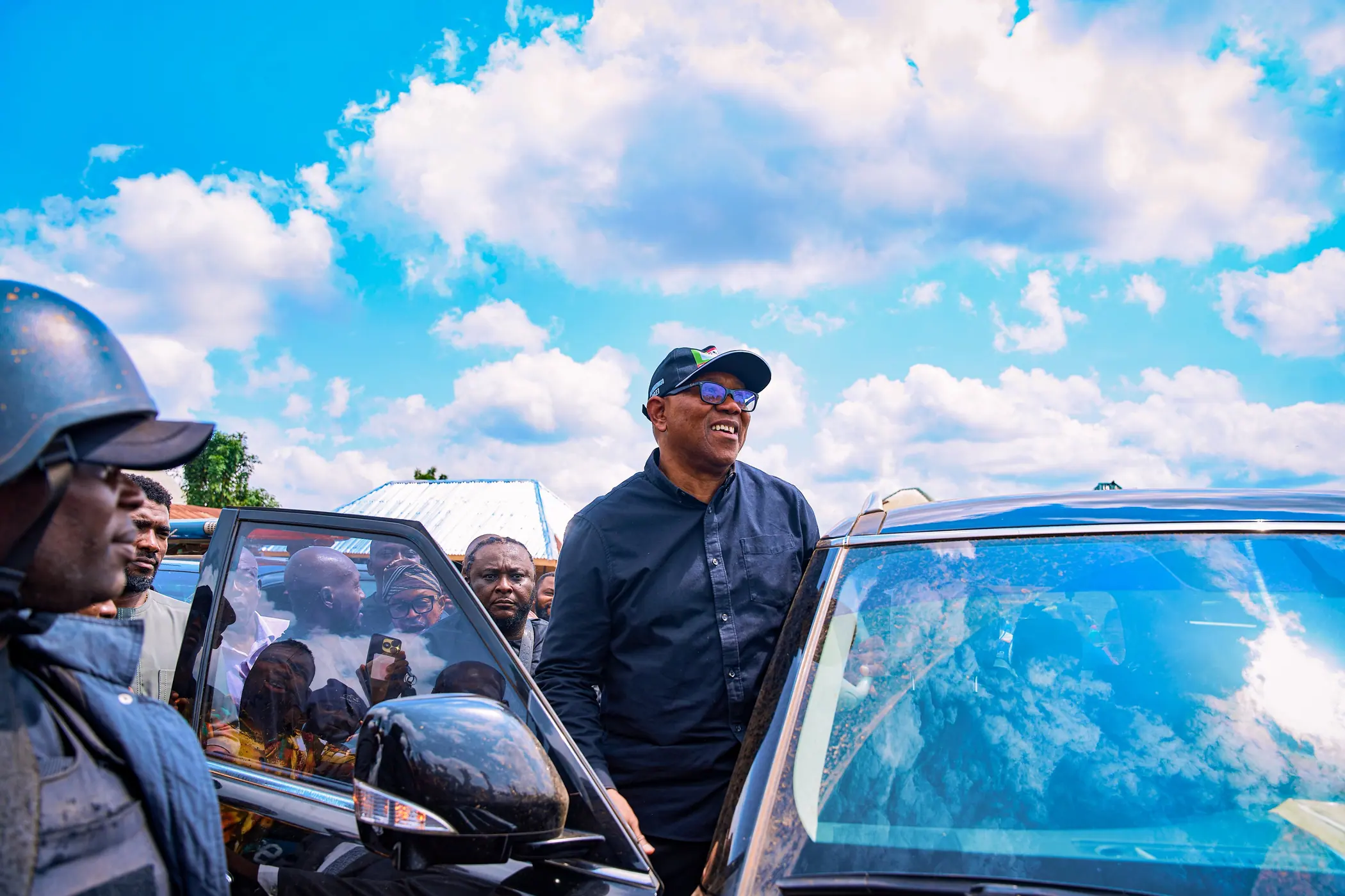 Peter Obi standing at the open door of a car under a bright sky, a crowd pressing in behind him
