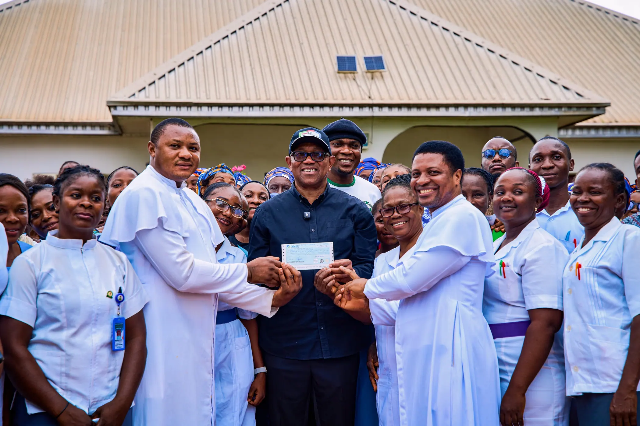 Peter Obi handing a cheque to staff outside a health facility, surrounded by nurses in white
