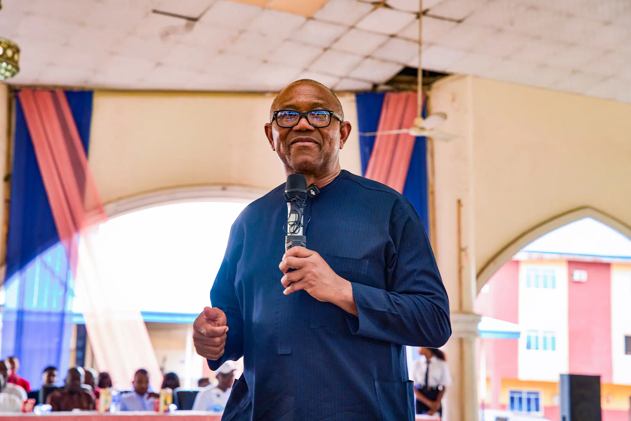 Peter Obi speaking into a handheld microphone under an arched hall roof
