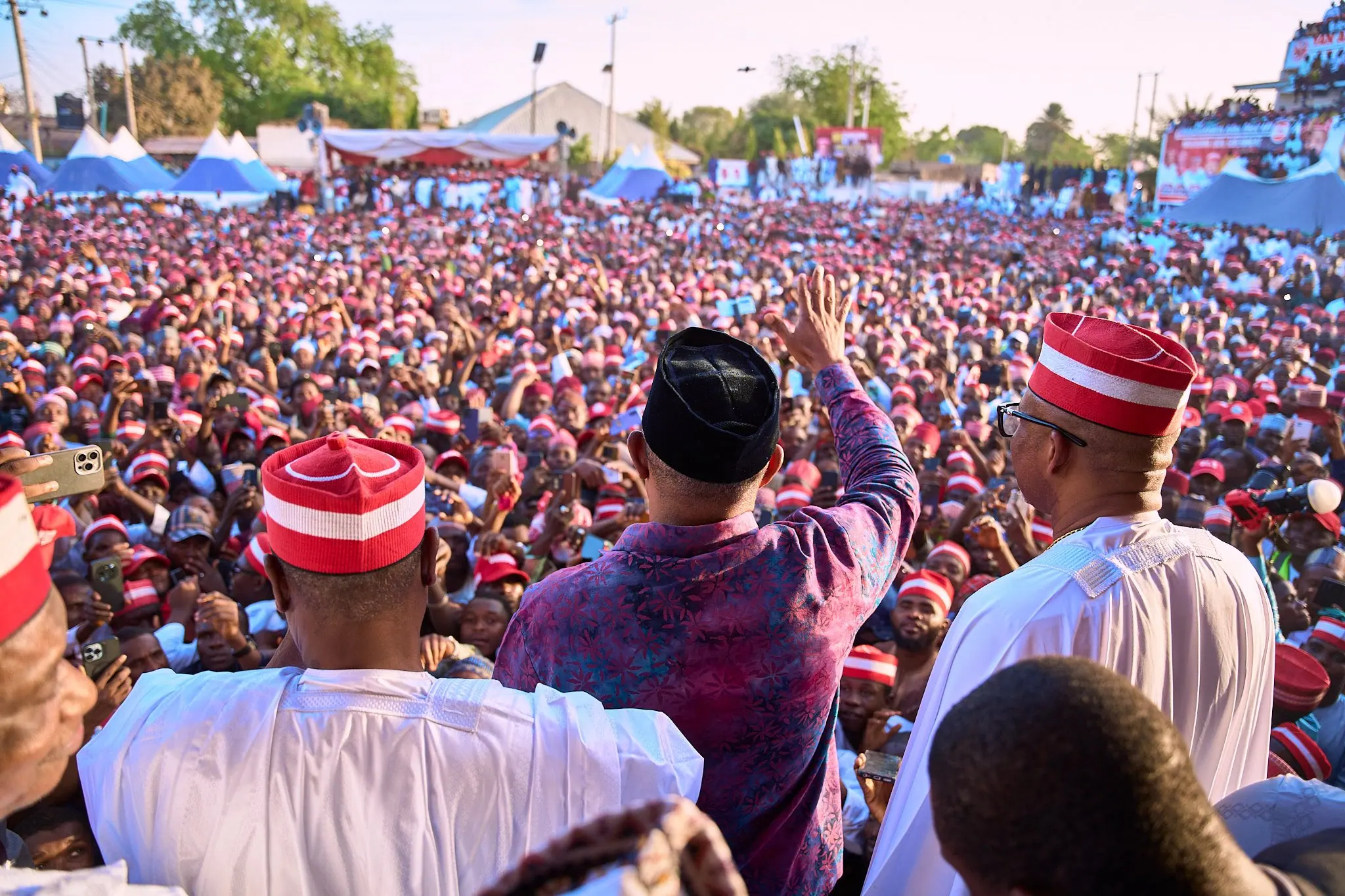 Rabiu Kwankwaso, Peter Obi and Bishop Isaac Idahosa photographed from behind on a platform, an arm raised to a crowd of tens of thousands in red and white Kano caps