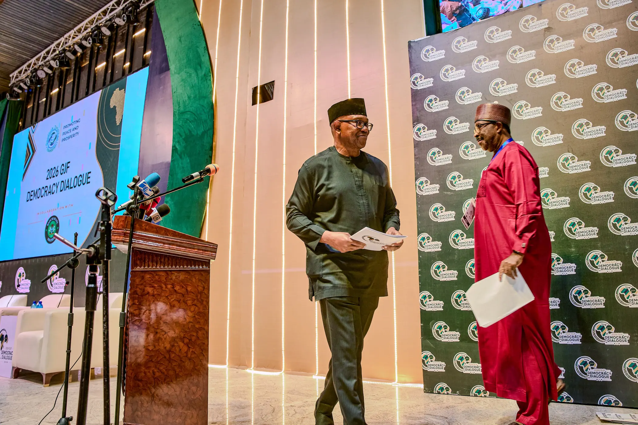 Peter Obi walking to a lectern at a conference stage, papers in hand, another delegate beside him