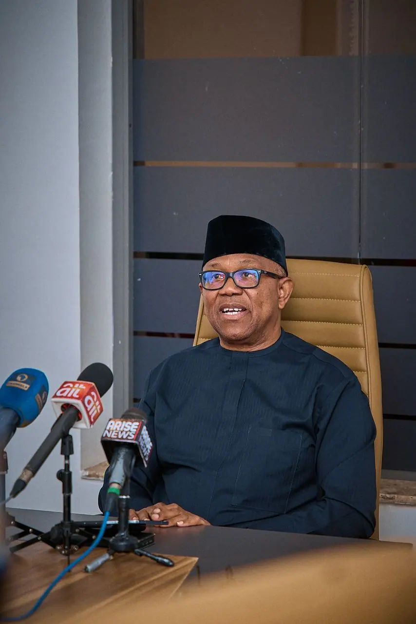 Peter Obi seated behind a desk in a black cap and navy tunic, facing a cluster of television microphones