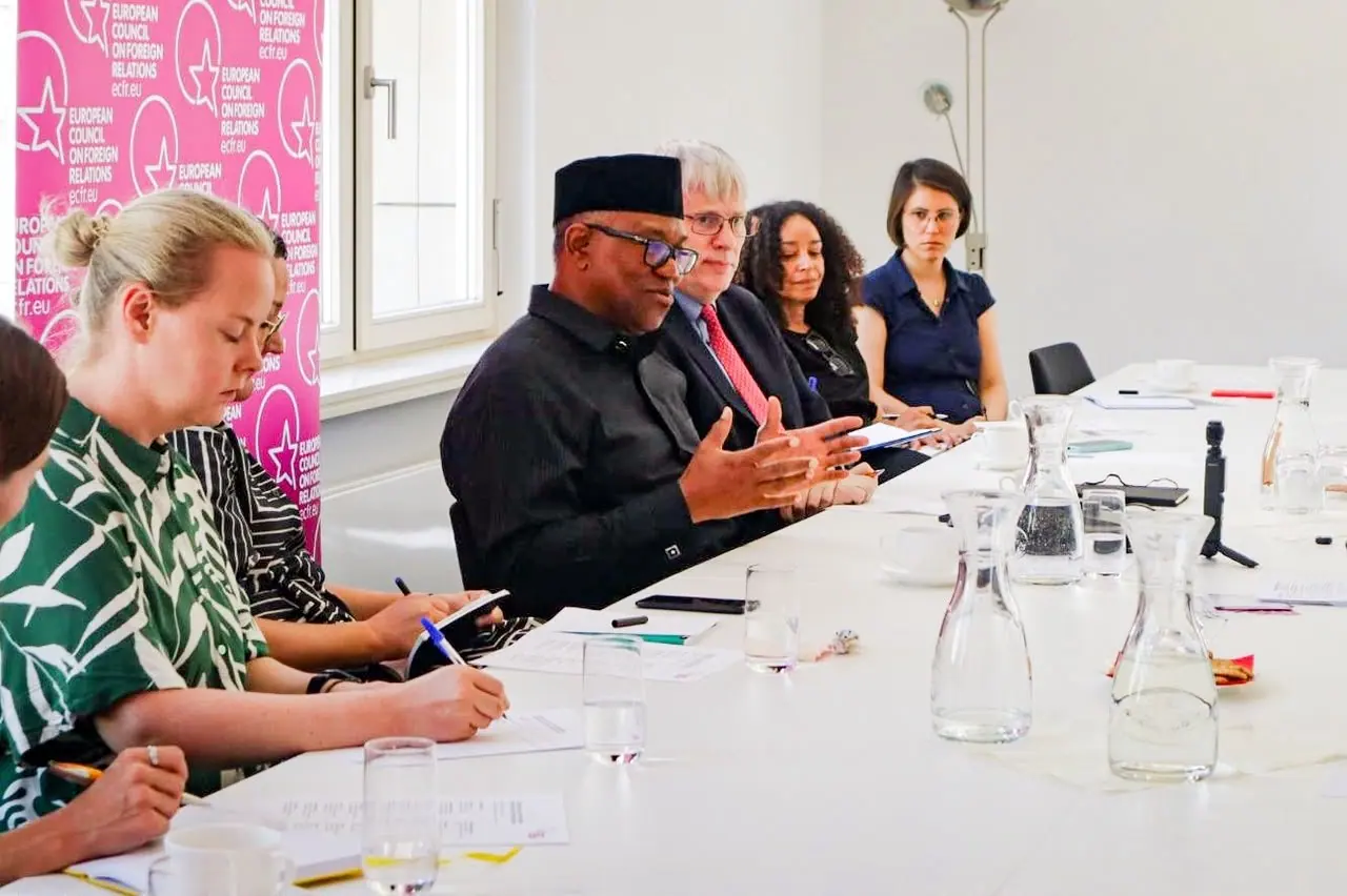 Peter Obi speaking with open hands at a long white table of researchers taking notes