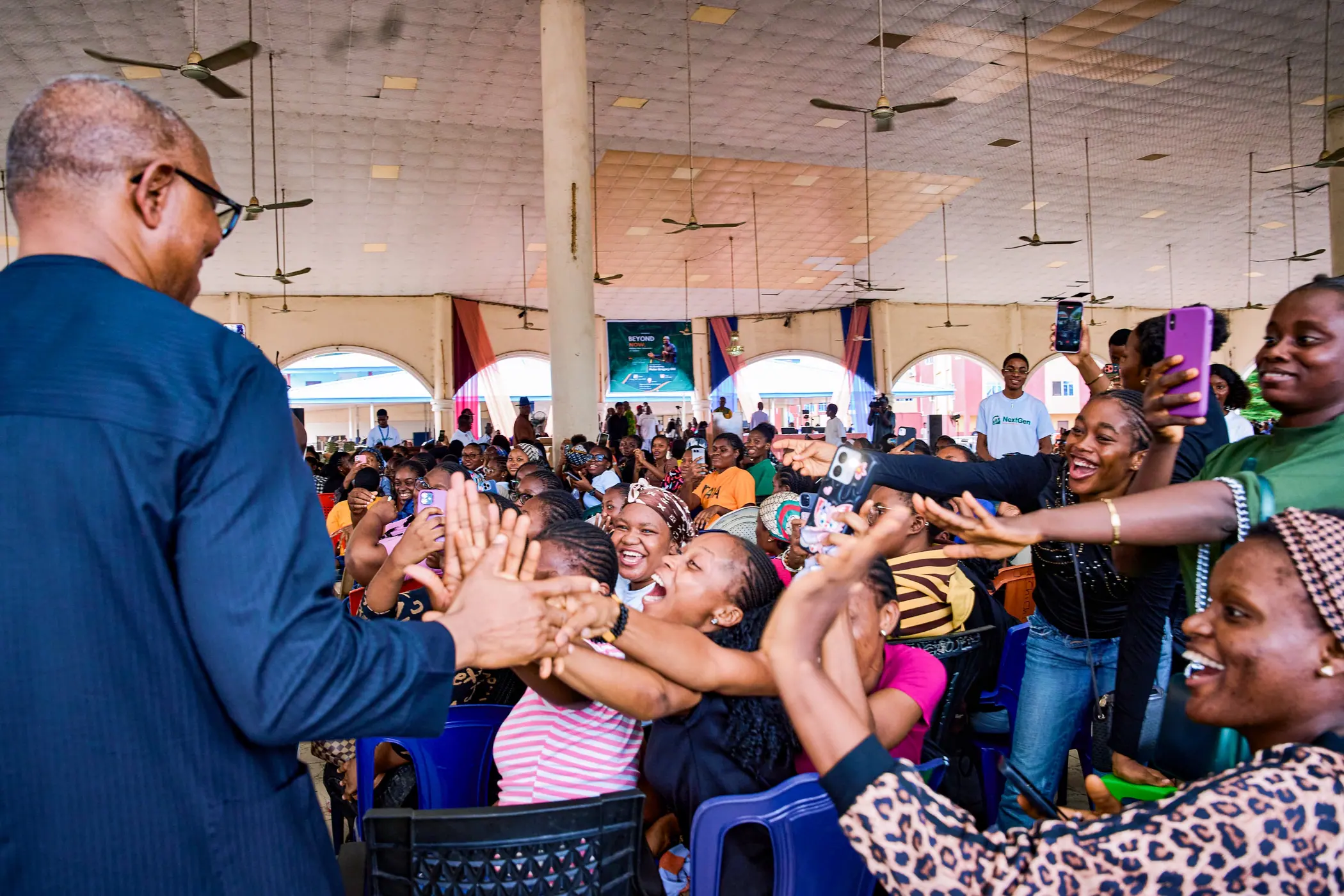 Peter Obi photographed from behind with his hand raised to a hall full of women and young people