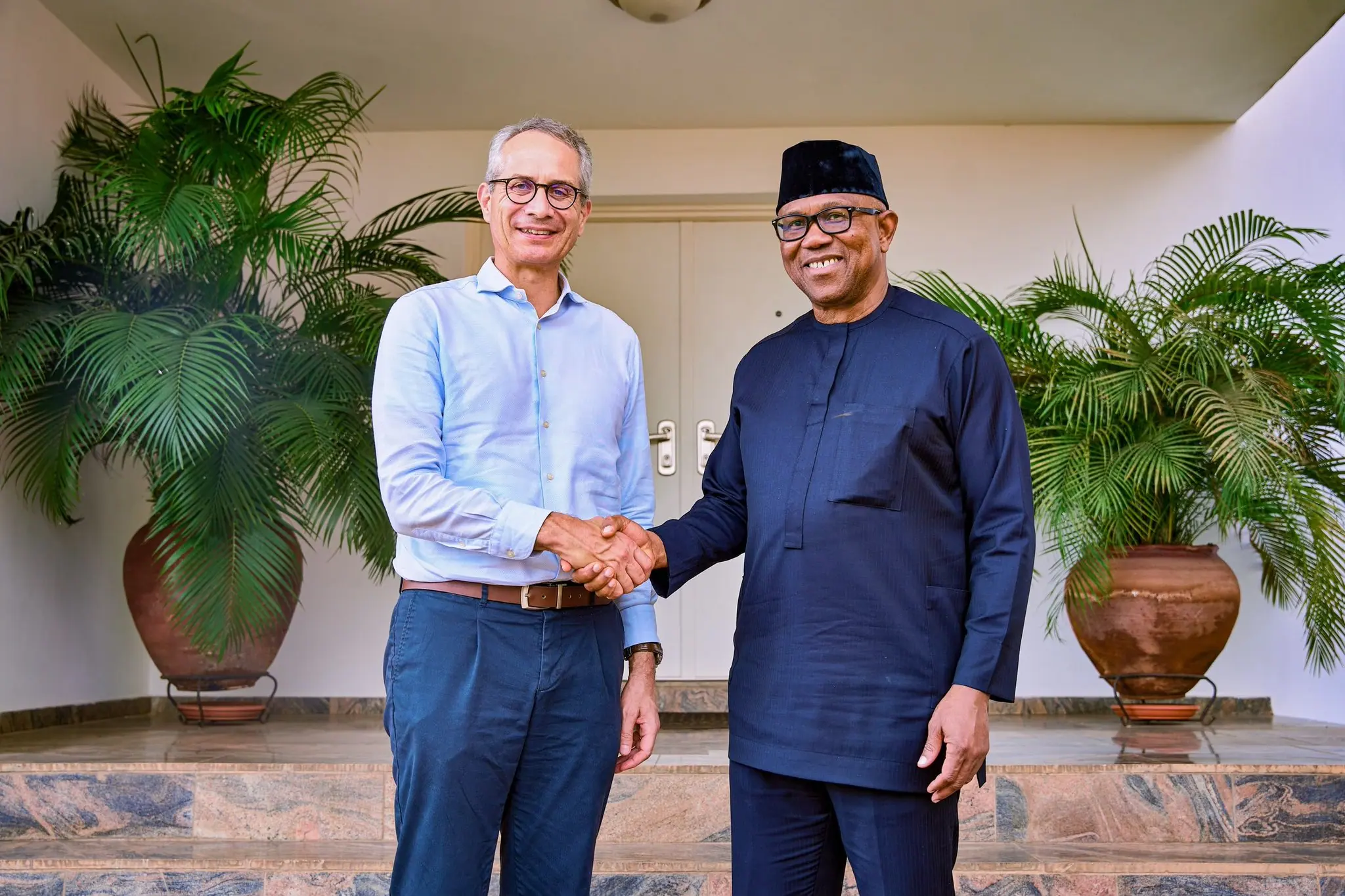 Peter Obi shaking hands with a visitor between two large potted palms