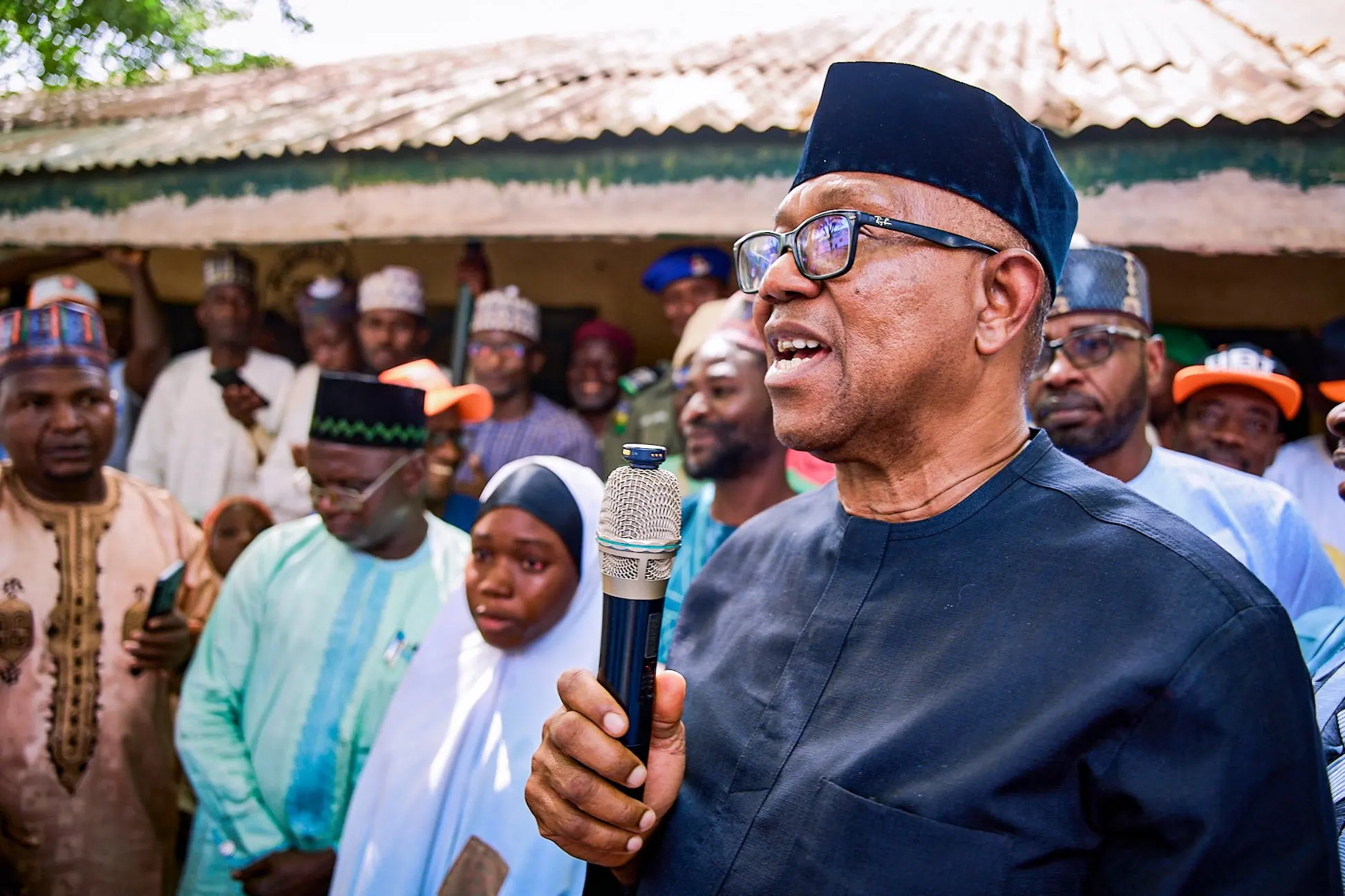 Peter Obi speaking into a microphone in a northern town, surrounded by men in caps and young women in hijabs