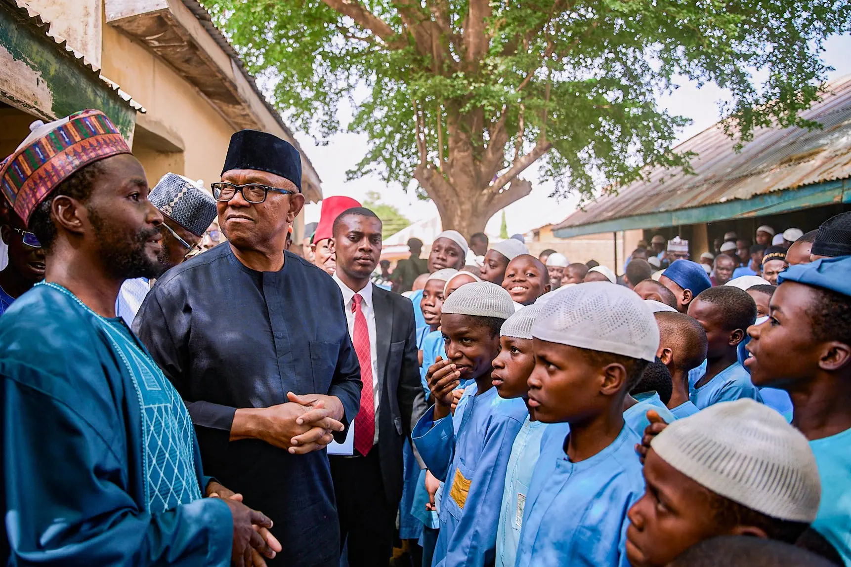 Peter Obi standing among schoolboys in blue uniforms and white caps under a tree in a northern town