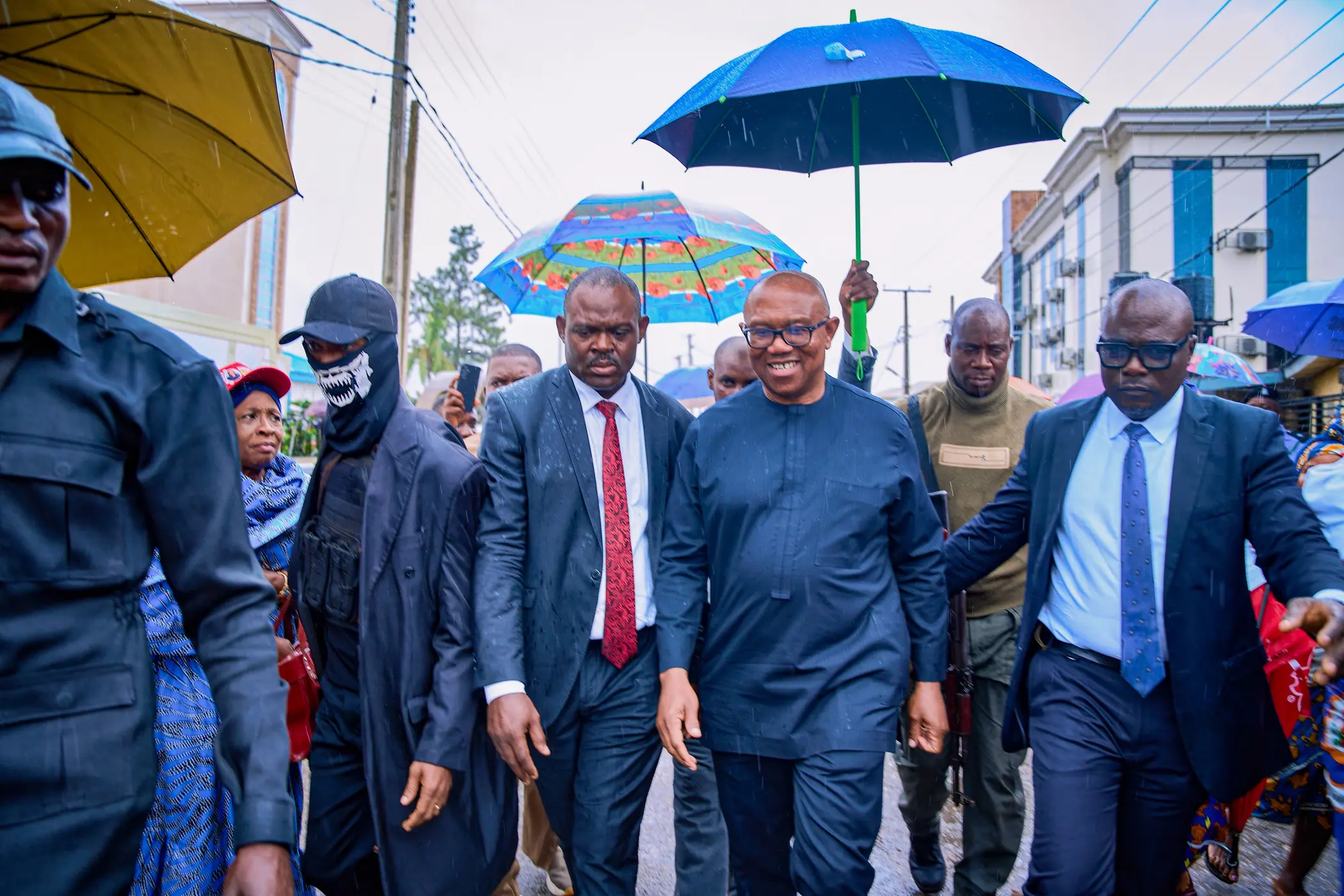 Peter Obi walking down a wet street with officials under blue and yellow umbrellas