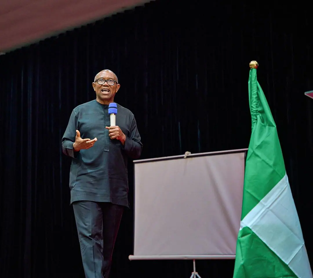 Peter Obi alone on a dark stage beside a Nigerian flag and a projection screen, microphone in hand