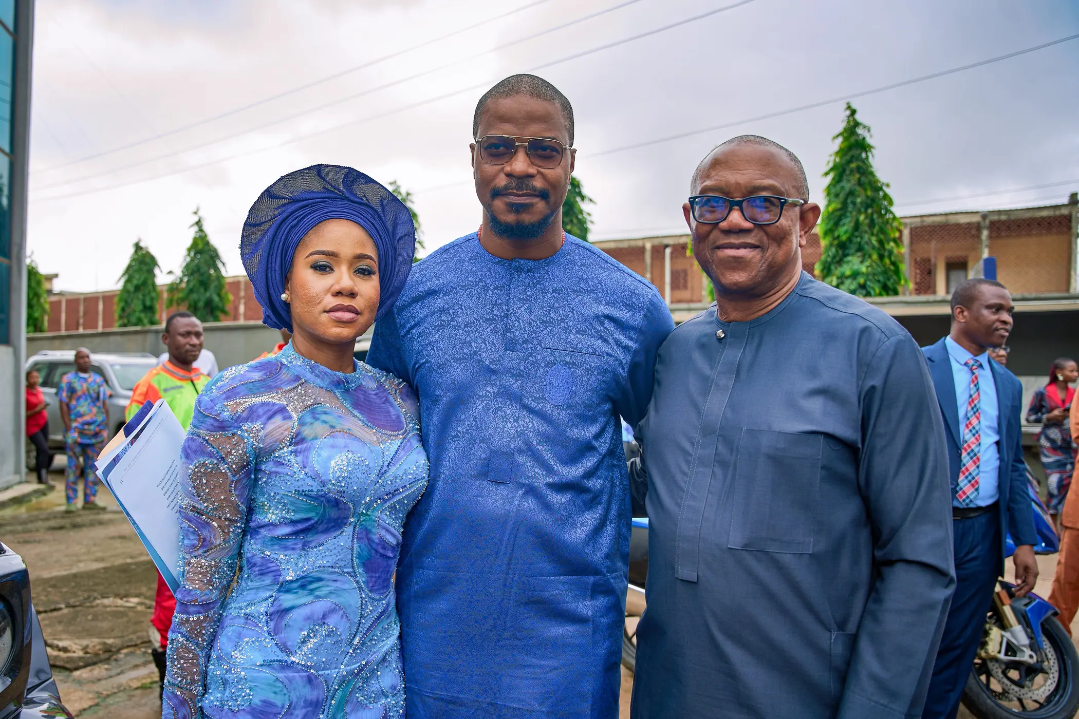 Peter Obi standing outside a building with a woman in a blue gele and a man holding a folder