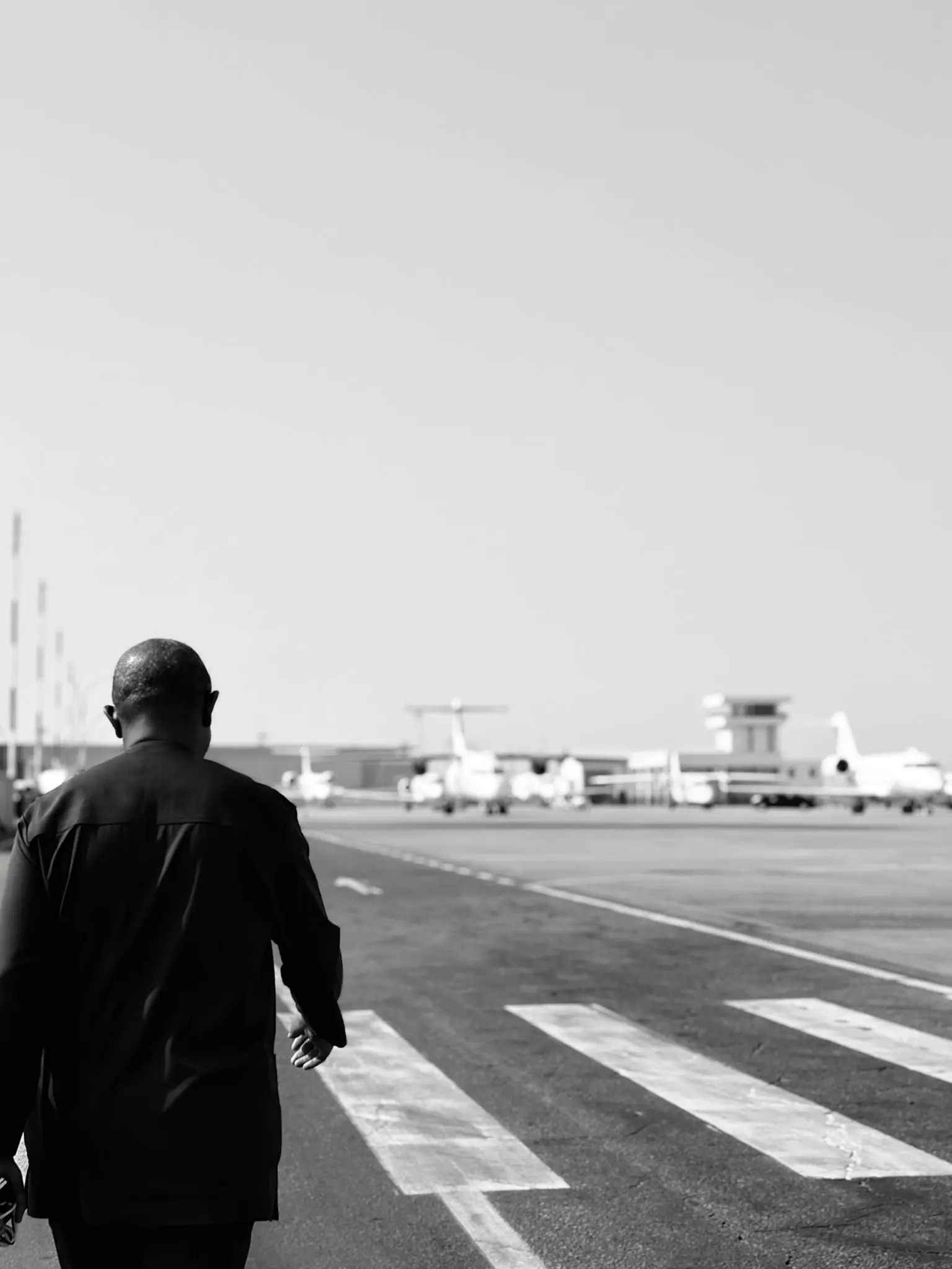 Black and white photograph of Peter Obi walking alone across an airport apron towards parked aircraft