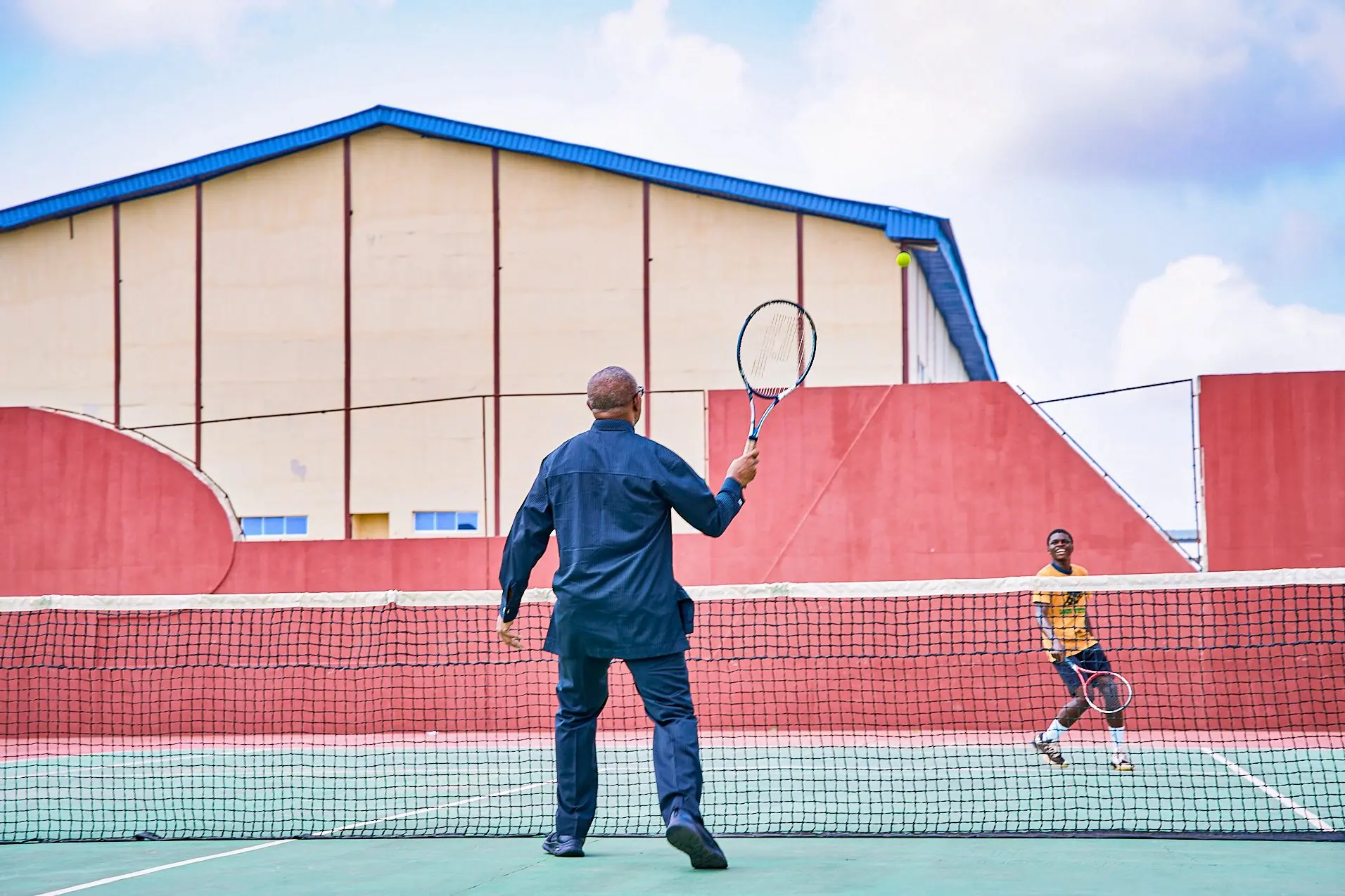 Peter Obi photographed from behind on a hard tennis court, racket raised, an opponent across the net