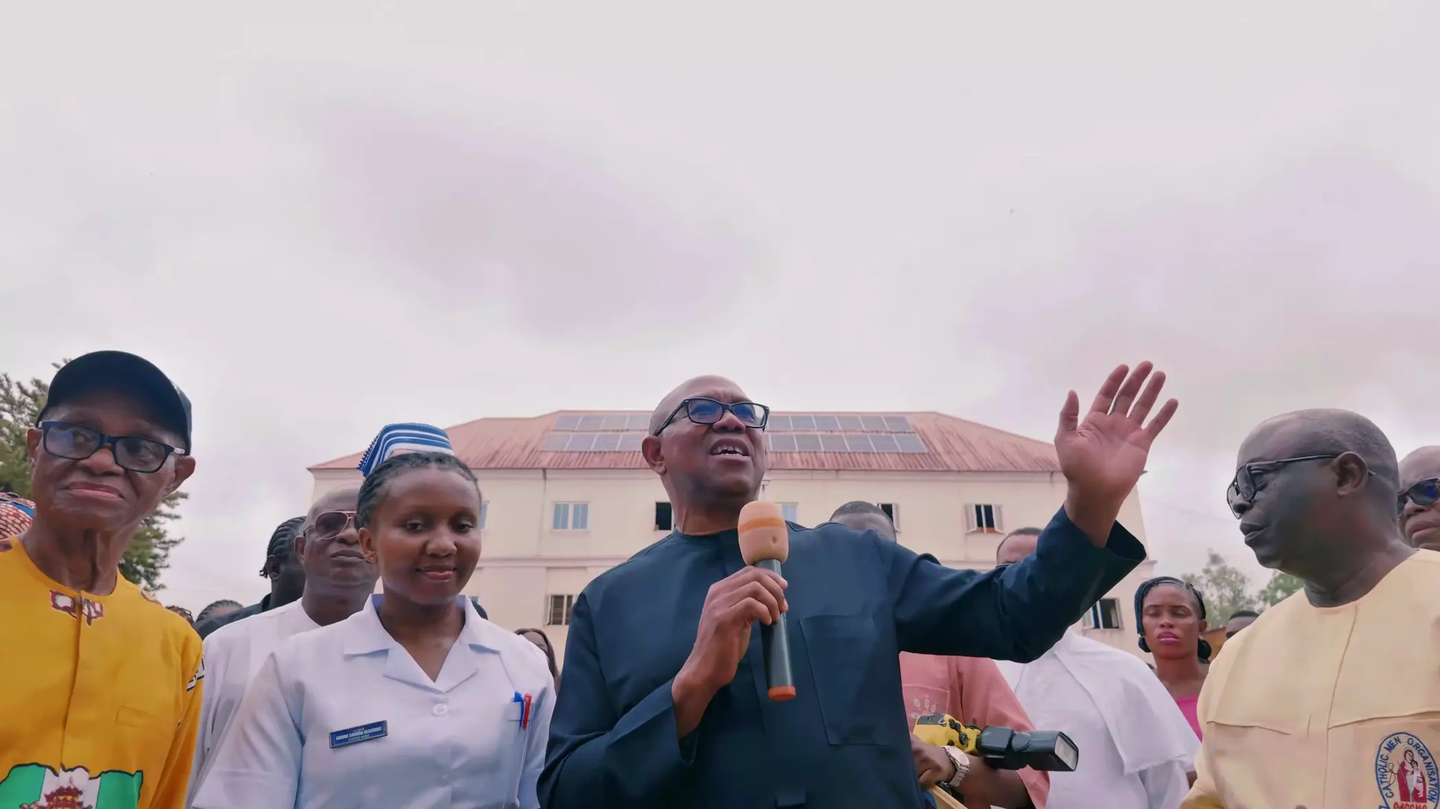 Peter Obi speaking with a microphone in a college courtyard, students and staff pressed around him